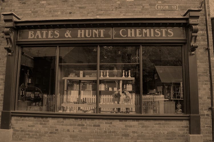 Sepia-tone picture of a store window, with a sign above saying “BATES & HUNTS CHEMISTS”