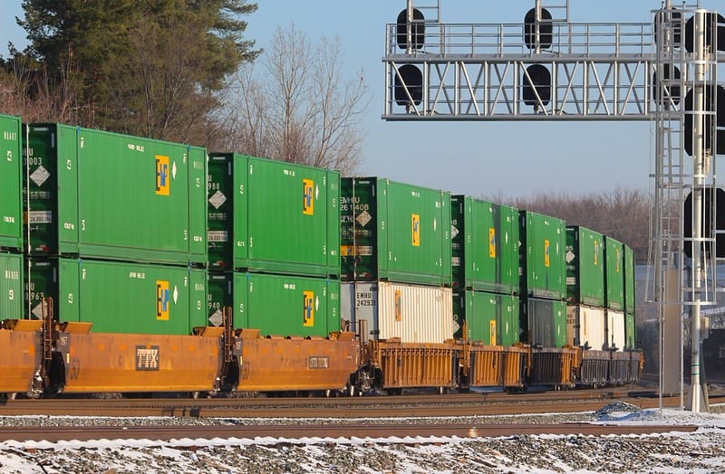 A large row of double-decker containers, mostly but not entirely green, on railcars curving into the distance.